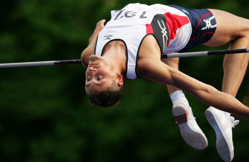 A high jumper arches over the bar mid-flight, his face locked in concentration as he executes the Fosbury Flop technique—the same mental discipline and pressure-tested composure that helps athletes outperform MBAs in tech companies during market crashes.