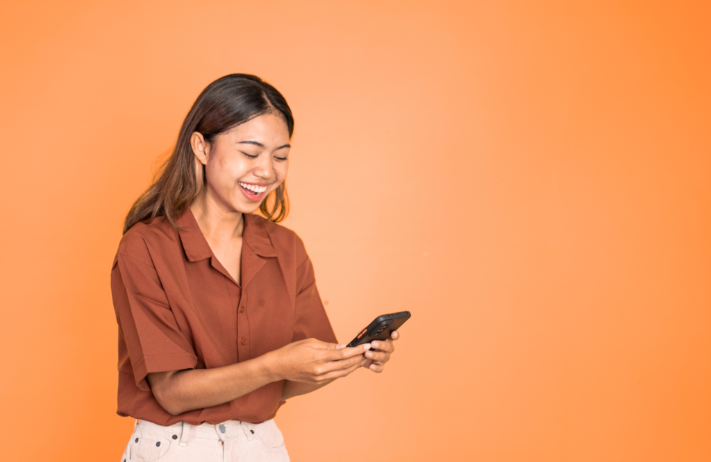 A smiling woman in a rust-colored button-up shirt looks down at her smartphone against a bright orange background, representing a content creator engaging with her audience or checking earnings.