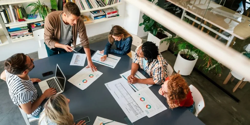 An overhead view of a marketing team gathered around a table covered in campaign analytics reports and performance charts, collaborating on influencer ROI strategy in a bright, modern workspace.